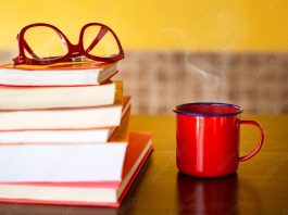 Pilha de livros com óculos vermelho em cima e uma caneca vermelha com café quente sobre a mesa.