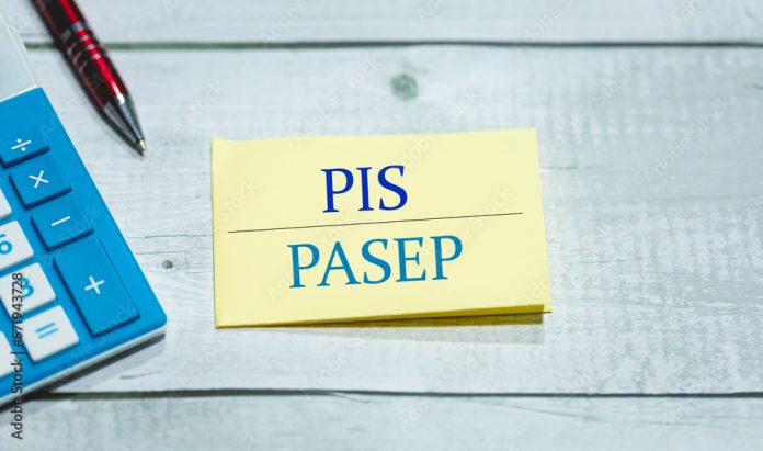 As siglas PIS e PASEP escritas em um pedaço de papel que está sobre uma mesa de madeira. Uma calculadora e uma caneta na composição. Economia brasileira.