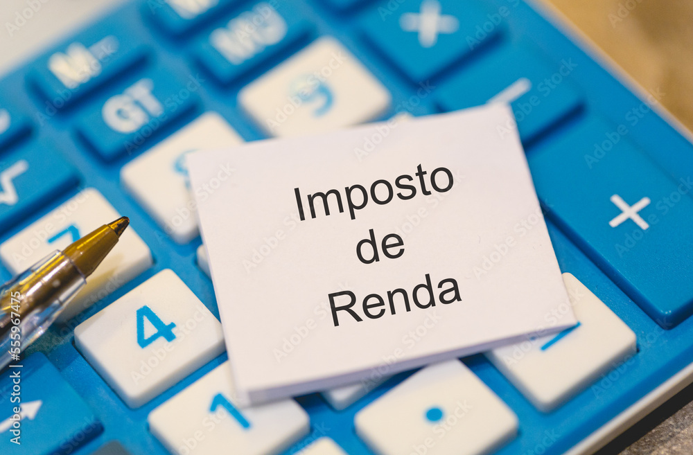 A palavra Imposto de Renda escrita em um pedaço de papel em língua Português do Brasil. Uma calculadora azul e uma caneta na composição. Economia brasileira.
