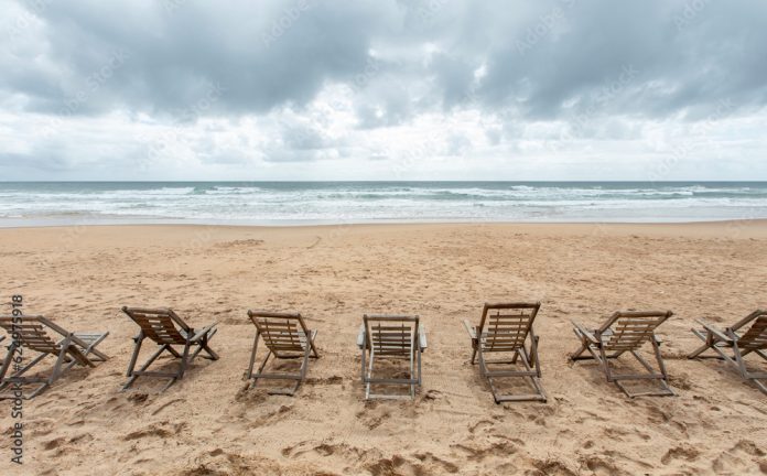 Espreguiçadeiras de madeira vazias na areia da praia deserta de frente para o mar em dia nublado.