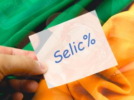 Selic. A palavra SELIC escrita em um papel de anotações de cor branca que está sendo segurado por um homem. Fundo com o Real, moeda brasileira e cores verde e amarelo. Economia, dinheiro, juros.