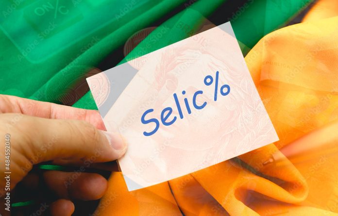 Selic. A palavra SELIC escrita em um papel de anotações de cor branca que está sendo segurado por um homem. Fundo com o Real, moeda brasileira e cores verde e amarelo. Economia, dinheiro, juros.
